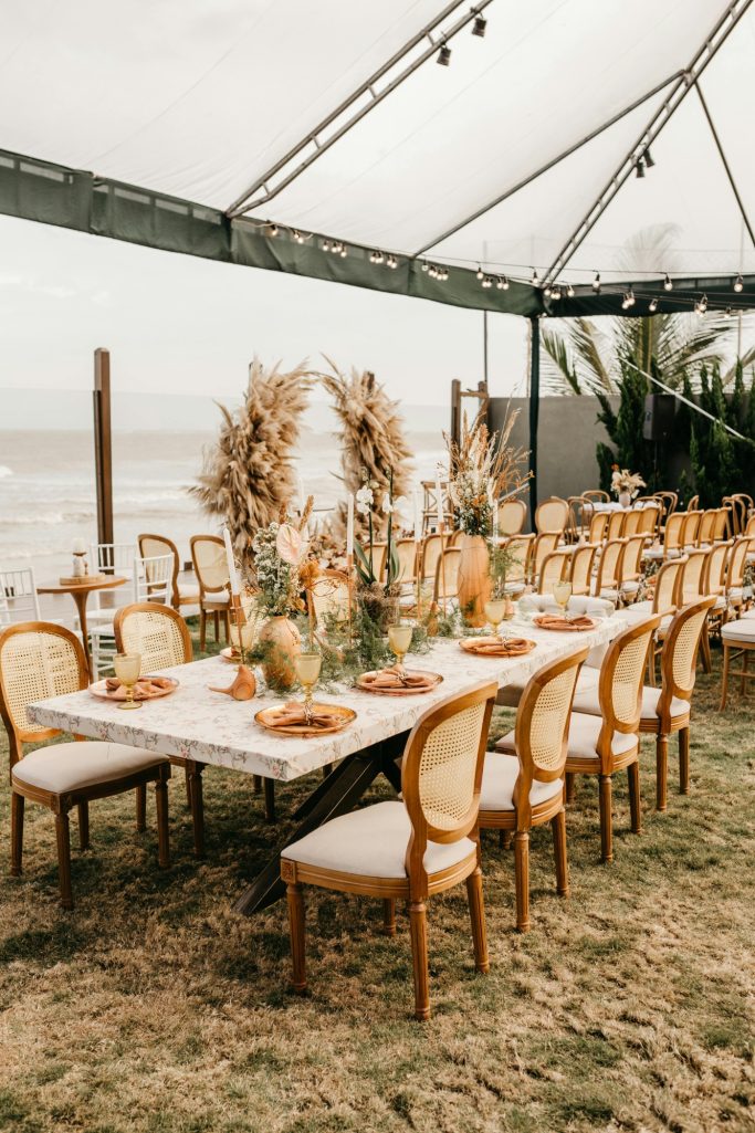 Elegant outdoor dining setup with a long table, chairs, and floral centerpieces near the beach.