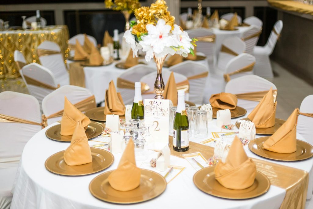 Elegant round dining tables set with gold and cream decor, featuring folded napkins and wine bottles.